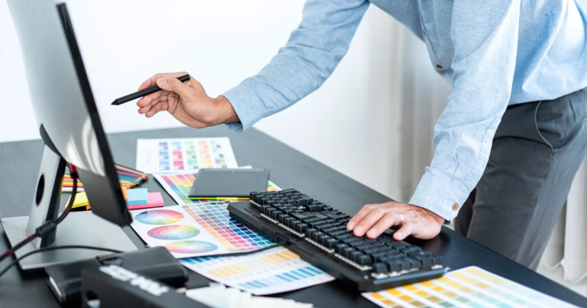 Person using a stylus to design with colour swatches on a desk beside a computer monitor.